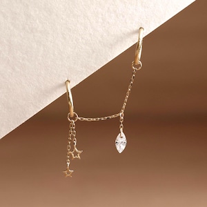 May include: A gold double hoop earring connected by a delicate chain. One hoop features two dangling open star charms, while the other hoop is linked to a single marquise-cut clear gemstone charm. The piece is crafted from polished yellow gold metal.