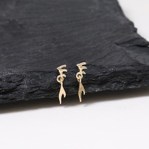 May include: A pair of gold-colored earrings with a unique, abstract design. Each earring features a small, decorative top element connected to a dangling, curved shape. The earrings are displayed on a dark, textured surface.