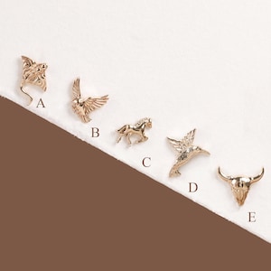May include: Five gold-toned metal charms labeled A through E. A is a stingray, B is a bird in flight, C is a horse, D is a hummingbird, and E is a bull skull. Each charm features detailed engravings and a polished finish on a white surface.