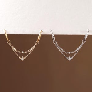 May include: Two double-chain hoop earrings, one in gold-tone and one in silver-tone metal. Each earring features two delicate chains connecting two hoops, with a small star-shaped charm containing a clear stone at the center of the lower chain.