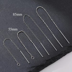 Peut inclure: Quatre boucles d'oreilles pendantes argentées de différentes longueurs. Deux paires sont étiquetées 55 mm et 85 mm. Les boucles d'oreilles comportent une chaîne de petites perles menant à une barre droite. Les boucles d'oreilles sont présentées sur une surface gris foncé.