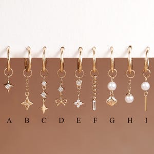Peut inclure: Neuf boucles d'oreilles créoles dorées étiquetées de A à I, chacune dotée d'une breloque pendante unique, notamment des étoiles, un nœud, une fleur, une chaîne, un coquillage, des perles et une barre, certaines ornées de petites pierres claires.