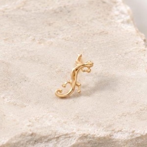 May include: A small gold-toned earring shaped like a lizard or gecko. The piece features a curved body with visible limbs and a tail, designed with a polished metallic finish. The earring is positioned to show its detailed, slender form.