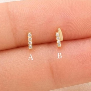 Peut inclure: Deux boucles d'oreilles à tige en or avec des pierres claires. L'option A est une barre verticale avec quatre petites pierres. L'option B est une barre légèrement inclinée avec trois pierres. Les deux bijoux sont petits et métalliques.