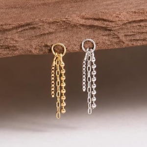 May include: Two dangle earrings, one gold-toned and one silver-toned, each featuring a circular hoop top with three delicate hanging chains. The gold earring has a mix of link and ball chains, while the silver earring features matching chain styles.