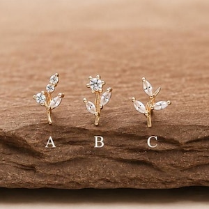 May include: Three gold-toned stud earrings shaped like botanical sprigs, each featuring clear, faceted stones. The earrings are labelled A, B, and C. Design A has three stones, design B has a central flower, and design C has three leaves.