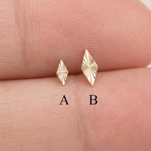 Puede incluir: Dos pequeños pendientes dorados en forma de diamante con textura de rayos de sol. La pieza marcada con A es más pequeña que la marcada con B. Ambos presentan un diseño geométrico puntiagudo con finas líneas grabadas desde el centro.