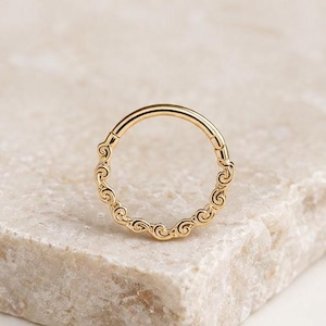 Può includere: Un anello circolare in oro caratterizzato da un motivo decorativo continuo di piccole spirali interconnesse lungo il bordo esterno. Il metallo ha una finitura lucida, creando una superficie riflettente che mette in risalto il design complesso e ripetitivo.