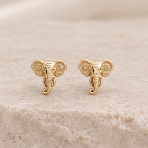 May include: A pair of gold stud earrings shaped like elephant heads. Each earring features a polished gold finish with engraved details on the ears and trunk, capturing the distinct facial features of an elephant in a small, symmetrical design.