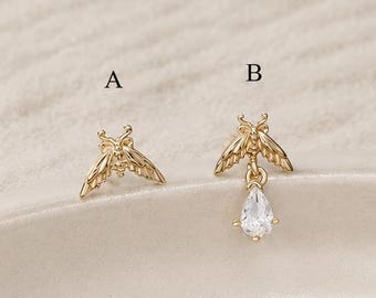 Könnte beinhalten: Zwei goldfarbene Ohrringe in Insektenform, beschriftet mit A und B. Ohrring A ist ein einfacher Ohrstecker. Ohrring B zeigt dasselbe Insektendesign mit einem hängenden, klaren, birnenförmigen Edelsteinanhänger. Beide Teile sind goldfarben.