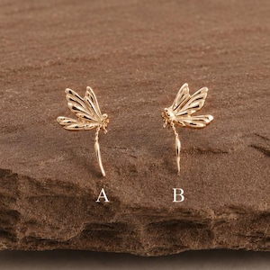 Peut inclure: Deux boucles d'oreilles à tige en forme de libellule dorée, étiquetées A et B, présentant des textures d'ailes détaillées et des corps fins. Chaque boucle d'oreille est un petit bijou délicat au fini métallique, arborant une forme d'insecte.