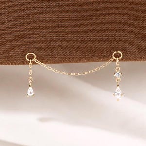 May include: A gold-toned chain earring featuring two circular hoops connected by a delicate link chain. Each hoop suspends a small vertical chain ending in a clear, pear-shaped gemstone drop, with an additional small round gemstone set above each drop.