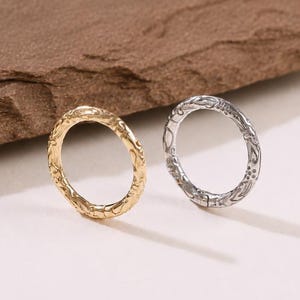 May include: Two circular rings, one in a gold tone and one in a silver tone, each featuring a delicate, repeating embossed pattern along the entire outer surface. The rings are positioned side by side, showcasing their thin, rounded metal bands.