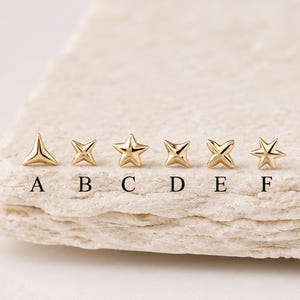 Op de afbeelding: Zes kleine gouden oorknopjes op een rij, gelabeld van A tot F. De ontwerpen bestaan uit een driehoek, een vierpuntige ster, een vijfpuntige ster, een ruitvorm, een kruisvormige ster en een zespuntige ster, allemaal met een gepolijste afwerking.