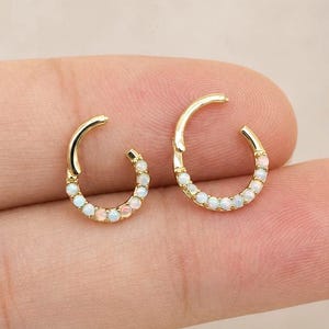 Op de afbeelding: Twee gouden oorringen met een scharnierend segmentontwerp. Elke ring is versierd met een rij kleine, iriserende witte opaalstenen langs de buitenste boog. De oorbellen hebben een gepolijste metalen afwerking en een ronde vorm.