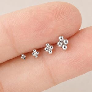 Könnte beinhalten: Vier silberfarbene Ohrstecker, die jeweils aus einer Anordnung von vier kleinen Metallkugeln in quadratischer Form bestehen. Die Ohrstecker sind in einer Reihe angeordnet, wobei die kleinste Größe links und die größte Größe rechts liegt.