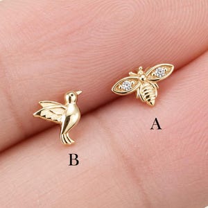 Puede incluir: Dos pendientes de metal en tono dorado. El pendiente A tiene forma de abeja con dos pequeñas piedras transparentes en las alas. El pendiente B tiene forma de pájaro en vuelo con las alas extendidas. Ambas piezas están marcadas con A y B.