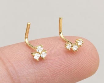 May include: Two gold nose studs, each featuring a trio of small, clear, round-cut stones arranged in a cluster. The studs have an L-shaped post design, with the metal finish appearing bright and polished against the light-colored background.