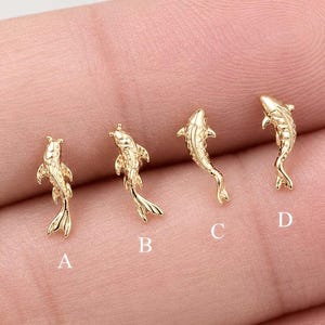 May include: Four small gold-toned koi fish charms or earring studs arranged in a row, labeled A, B, C, and D. Each piece features a detailed, textured scale pattern and a curved tail design, showcasing a metallic finish and a distinct swimming posture.