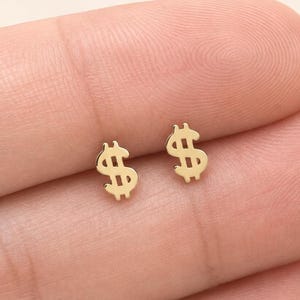 以下が含まれることがあります： ドル記号の形をした小さなゴールドトーンのスタッドピアス一対。各ピアスは光沢のある金属仕上げで、文字Sの中央に縦線が入ったシンプルでクリーンなデザインが特徴です。耳たぶの上に置かれた状態で撮影されています。