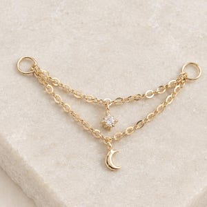 May include: A gold-toned double chain connector featuring two circular end loops. The upper chain holds a small, four-pointed star charm with a central clear stone, while the lower chain features a small, smooth crescent moon charm.