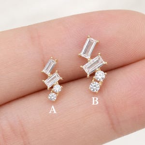 May include: Two gold-tone stud earrings labelled A and B. Earring A features two rectangular baguette-cut clear stones and two round clear stones. Earring B features two rectangular baguette-cut clear stones and two round clear stones in a larger cluster.