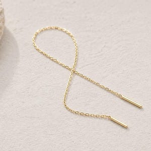 Könnte beinhalten: Ein goldener Durchziehohrring mit einer feinen, zarten Kette, die eine Unendlichkeitsform bildet und an beiden Enden mit zwei kleinen, glatten, zylindrischen Metallstäbchen versehen ist, was ein schlichtes und durchgehendes Design ergibt.