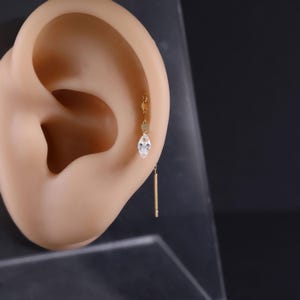 May include: A gold-coloured earring with a dangling design. The earring features a clear, teardrop-shaped gem and a small, vertical bar. The earring is shown on a mannequin ear against a dark background.