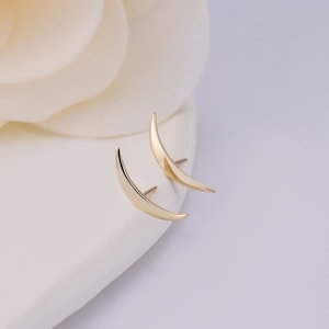 May include: A pair of gold-toned crescent moon stud earrings with a smooth, polished finish. Each earring features a curved, tapered design that comes to a point at both ends, with a thin metal post extending from the back for secure attachment.