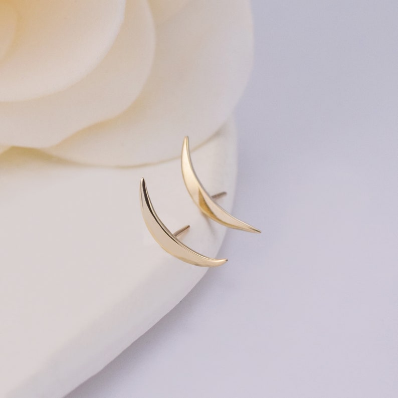May include: A pair of gold-toned crescent moon stud earrings with a smooth, polished finish. Each earring features a curved, tapered design that comes to a point at both ends, with a thin metal post extending from the back for secure attachment.