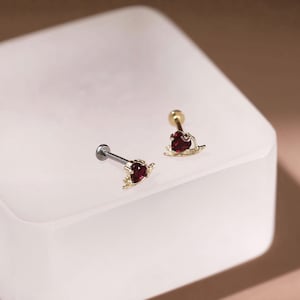 May include: Two labret stud earrings featuring a small, deep red heart-shaped gemstone set in gold-toned metal. Each stud has a flat back disc and a decorative base element resembling small leaves or wings beneath the heart-shaped stone.