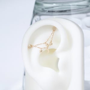 May include: A gold-toned ear cuff featuring two delicate chains connected to two small star charms and a crescent moon charm. The jewelry piece is designed to wrap around the upper cartilage of the ear, secured by two small circular studs at each end.