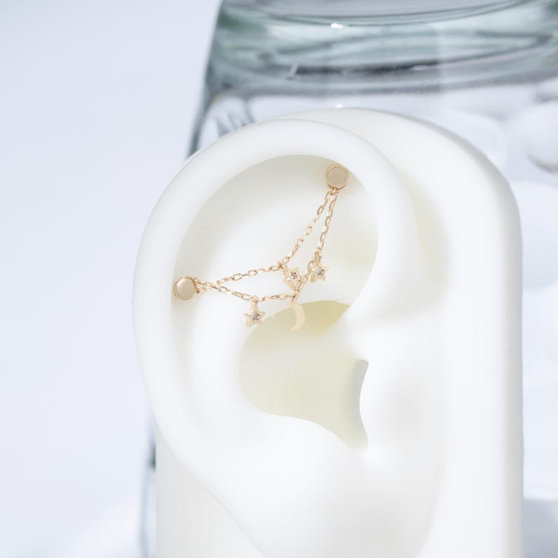 May include: A gold-toned ear cuff featuring two delicate chains connected to two small star charms and a crescent moon charm. The jewelry piece is designed to wrap around the upper cartilage of the ear, secured by two small circular studs at each end.