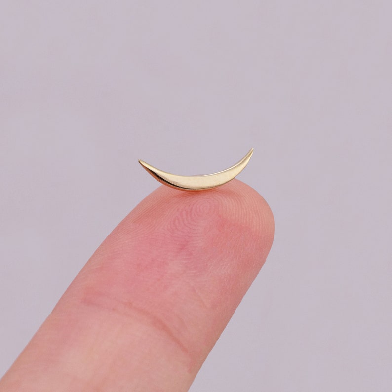 May include: A small, polished gold-colored earring stud shaped like a slender crescent moon. The piece features a smooth, reflective metallic surface with tapered, pointed ends, designed to sit flat against the skin when worn as a piece of jewelry.