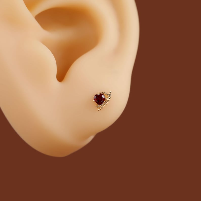 May include: A gold stud earring featuring a small, deep red heart-shaped gemstone. The setting includes a delicate, curved gold branch-like detail extending from one side of the heart, creating a subtle, asymmetrical design on the earlobe.