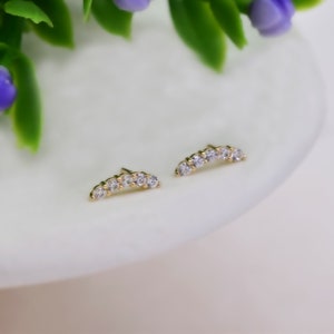 May include: A pair of gold stud earrings, each featuring a curved bar design set with five small, clear, round-cut stones. The earrings have a polished metallic finish and a delicate, arched shape that follows the contour of the earlobe.