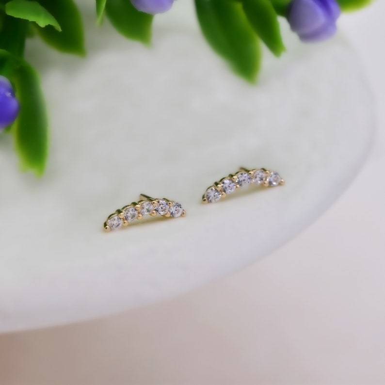 May include: A pair of gold stud earrings, each featuring a curved bar design set with five small, clear, round-cut stones. The earrings have a polished metallic finish and a delicate, arched shape that follows the contour of the earlobe.