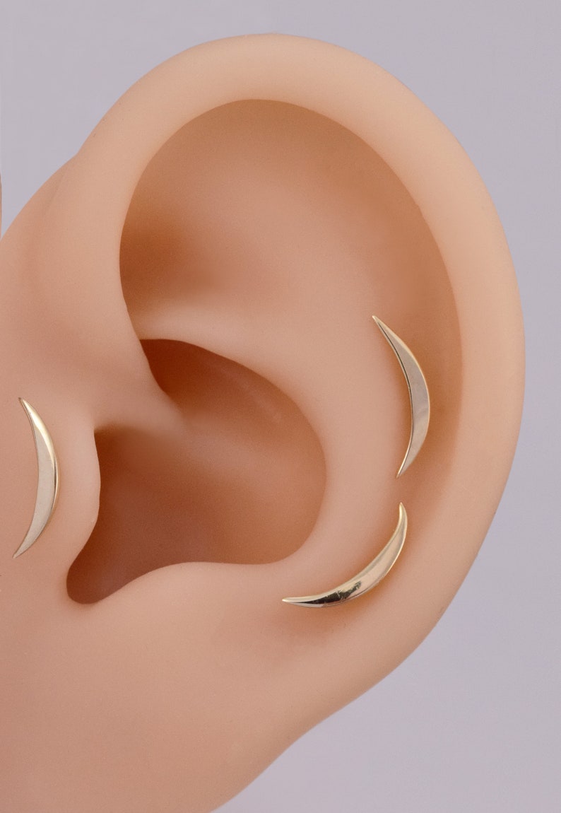 May include: Three gold-colored crescent moon-shaped earrings positioned on an ear model. Each earring features a smooth, curved, metallic surface that tapers to fine points at both ends, creating a sleek and simple design for ear piercings.