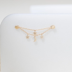 May include: A gold-toned double chain earring connector featuring two small circular studs at each end. The delicate chains are adorned with three dangling charms: two five-pointed stars and a central crescent moon, each with a small clear stone.