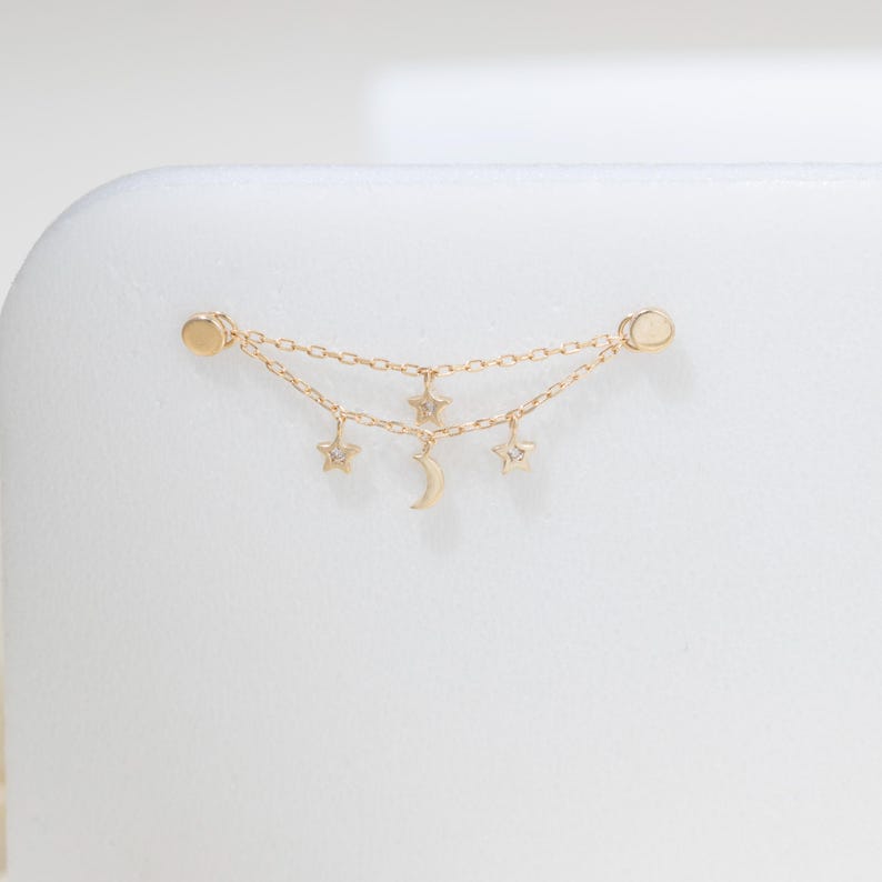 May include: A gold-toned double chain earring connector featuring two small circular studs at each end. The delicate chains are adorned with three dangling charms: two five-pointed stars and a central crescent moon, each with a small clear stone.
