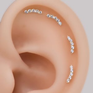 May include: Four gold-toned curved ear piercings arranged along the cartilage of an ear. Each piece features a row of five small, clear, round-cut stones set in a metal arc. The jewelry is positioned in a vertical and diagonal pattern.