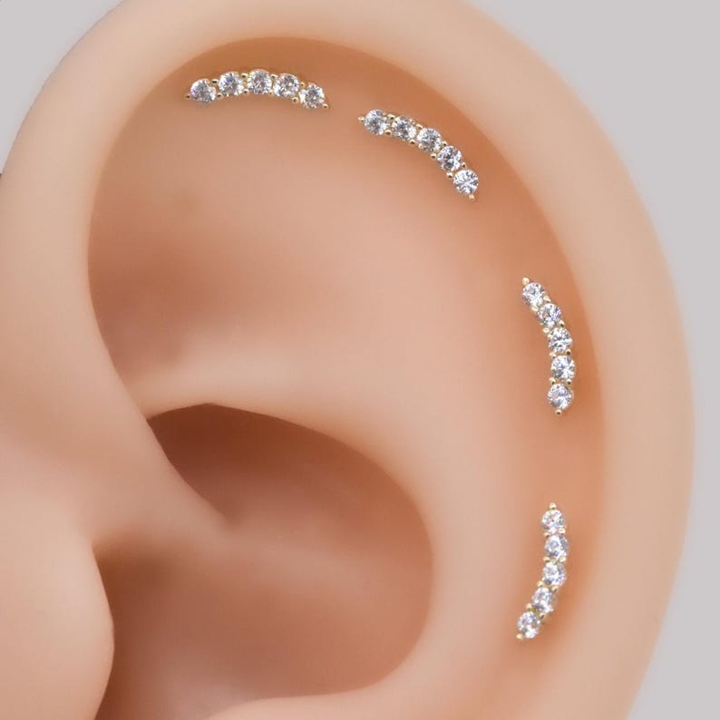 May include: Four gold-toned curved ear piercings arranged along the cartilage of an ear. Each piece features a row of five small, clear, round-cut stones set in a metal arc. The jewelry is positioned in a vertical and diagonal pattern.
