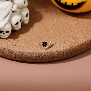 May include: A small silver stud earring featuring a dark red heart-shaped gemstone. The earring has a metallic setting that holds the stone securely in place, showcasing a polished finish and a deep crimson color in the center of the heart.