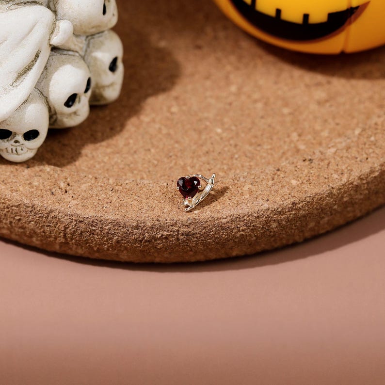 May include: A small silver stud earring featuring a dark red heart-shaped gemstone. The earring has a metallic setting that holds the stone securely in place, showcasing a polished finish and a deep crimson color in the center of the heart.