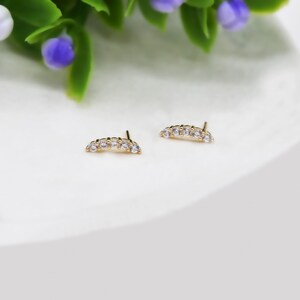 May include: A pair of gold stud earrings, each featuring a curved bar design set with five small, clear, round-cut stones. The earrings have a post back closure and are positioned side by side, showcasing a polished metallic finish and clear stones.