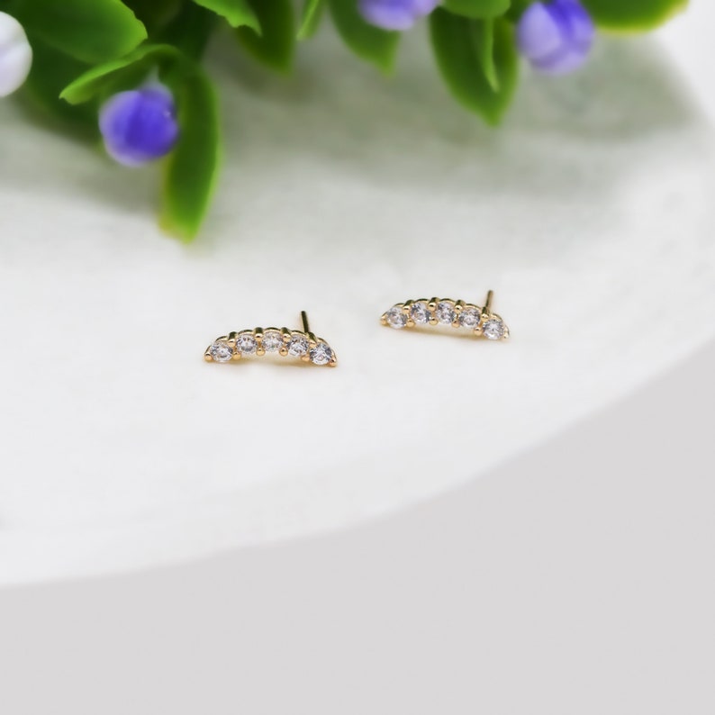 May include: A pair of gold stud earrings, each featuring a curved bar design set with five small, clear, round-cut stones. The earrings have a post back closure and are positioned side by side, showcasing a polished metallic finish and clear stones.