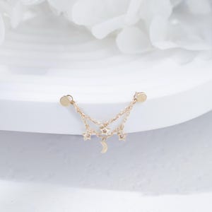 May include: A gold-toned earring featuring two delicate chains connected to two circular studs. The chains are adorned with three small star-shaped charms and one crescent moon charm hanging from the center, creating a layered celestial design.