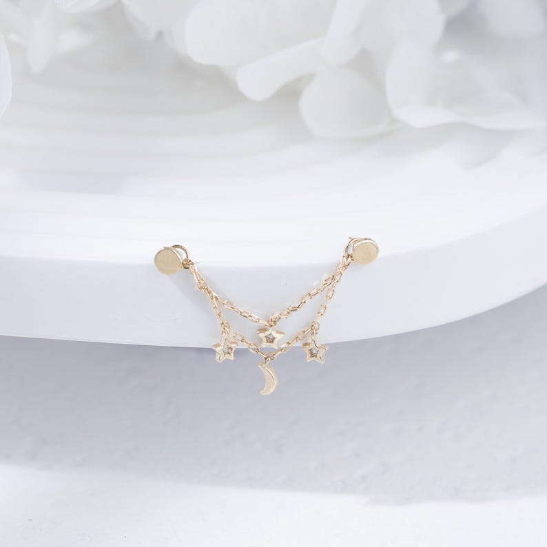 May include: A gold-toned earring featuring two delicate chains connected to two circular studs. The chains are adorned with three small star-shaped charms and one crescent moon charm hanging from the center, creating a layered celestial design.
