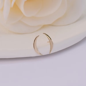 May include: A gold-toned earring featuring a curved, crescent-shaped design that mimics the appearance of two opposing horns or a split moon. The metal surface is smooth and polished, reflecting light along its tapered, pointed ends.