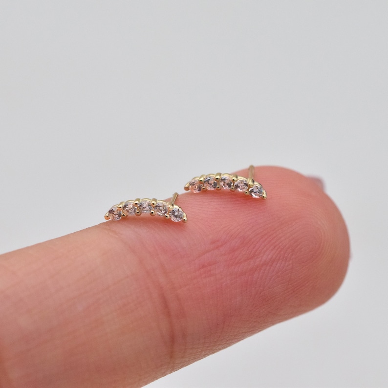 May include: A pair of gold stud earrings, each featuring a curved bar design set with five small, clear, round-cut stones. The earrings are crafted with a polished gold finish and include a post backing for secure attachment to the earlobe.
